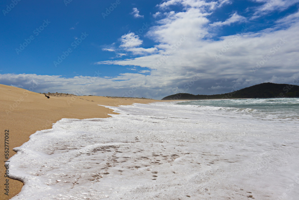 Waves crashing at Fingal Beach with people walking along the sand in the distance on a sunny day. Waves crashing at Fingal Beach with people walking along the sand in the distance on a sunny day.
