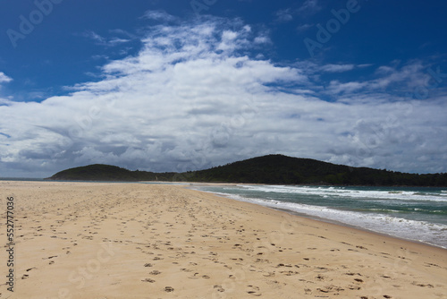 Waves crashing at Fingal Beach with people walking along the sand in the distance on a sunny day.