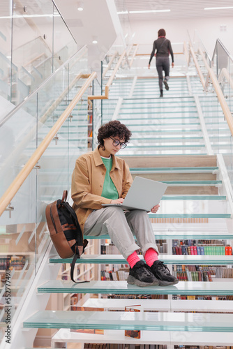 woman with curly hair is sitting on steps and working on laptop. Spanish female student or freelancer in library or coworking. thoughtful business person watch training webinar