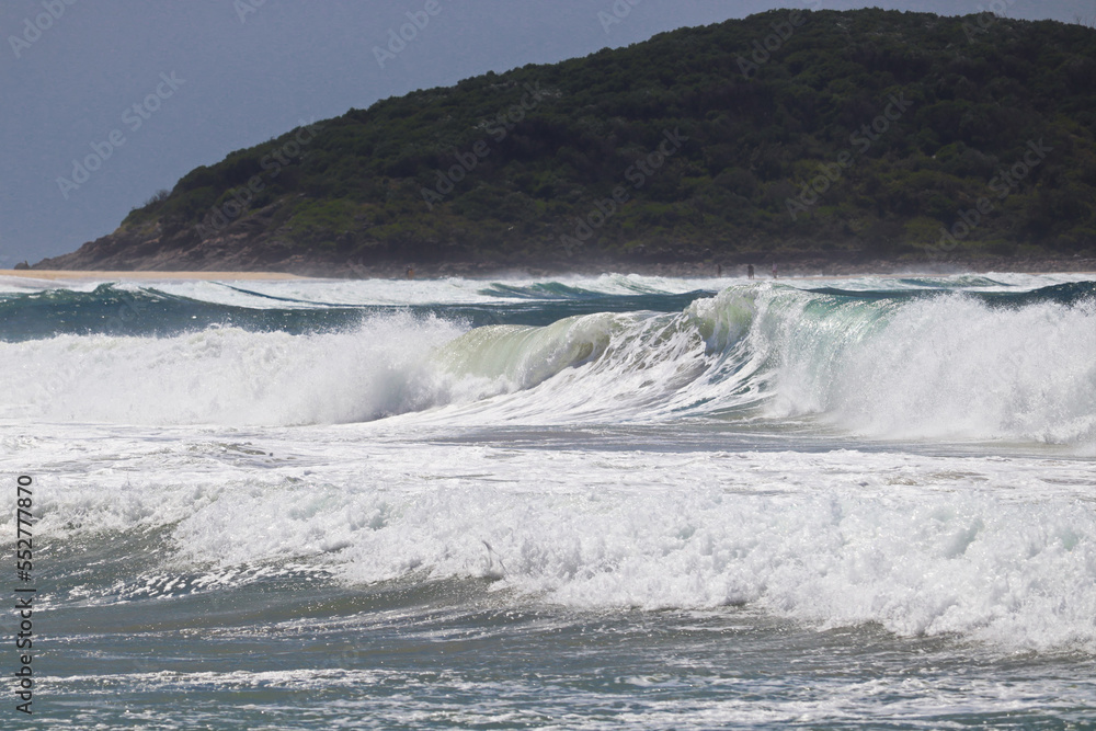 Waves crashing at Fingal Beach with people walking along the sand in the distance on a sunny day.