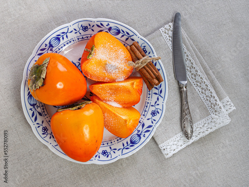 Whole and halved ripe persimmon fruits on a plate standing on a laid table, a table knife lies nearby