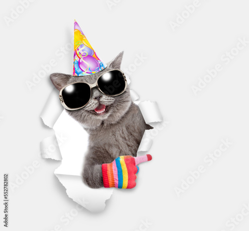 Happy cat wearing sunglasses and birthday cap looks through a hole in white paper and points away on empty space