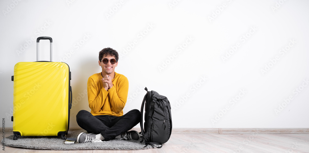 Young guy traveler with a suitcase satisfied with a positive expression sits isolated on a white background holds his hands near his chest Young guy traveler with a suitcase satisfied with a positive expression sits isolated on a white background holds his hands near his chest