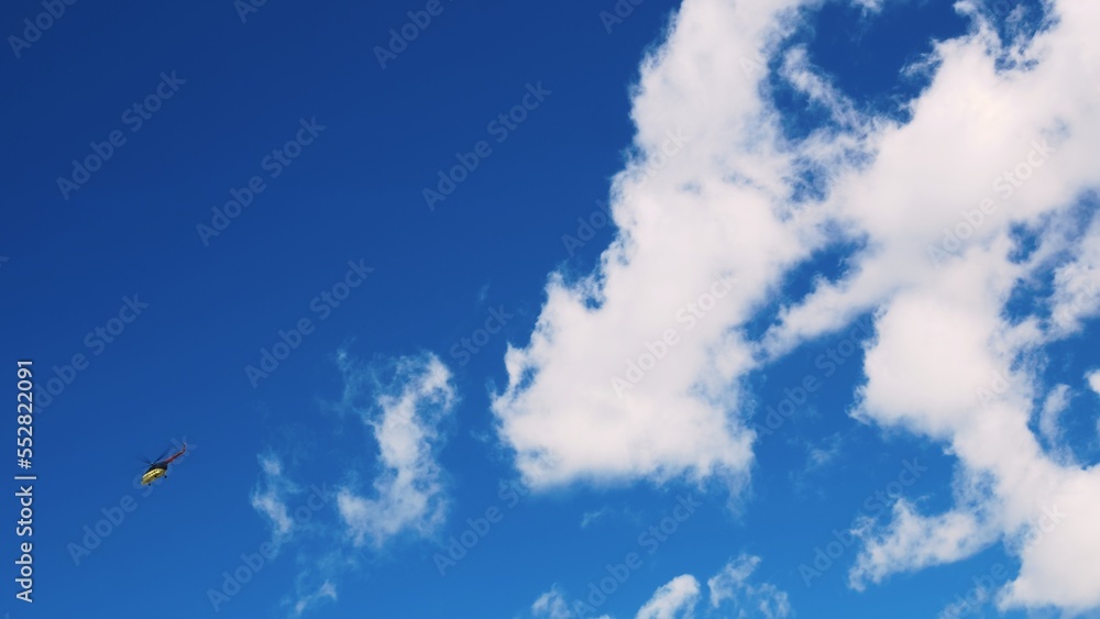 The blue sky is densely covered with cumulus clouds. The camera moves from right to left, following a yellow helicopter flying in the distance. A yellow helicopter flies through the cloudy sky. The blue sky is densely covered with cumulus clouds. The camera moves from right to left, following a yellow helicopter flying in the distance. A yellow helicopter flies through the cloudy sky.