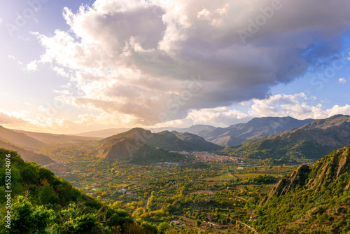 beautiful landscape with amazing view from highland with green slopes and bushes to a valley with town, road and nice mountains and scenic cloudy sunset on background