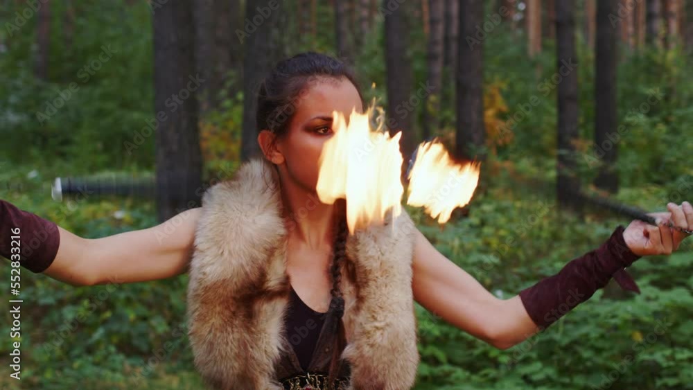 Girl in dress for performance spins ignited torches over her head and around her in boreal forest. Siberian female Shaman conjures. Woman participates in fire show in wooded place. Magic juggling Girl in dress for performance spins ignited torches over her head and around her in boreal forest. Siberian female Shaman conjures. Woman participates in fire show in wooded place. Magic juggling