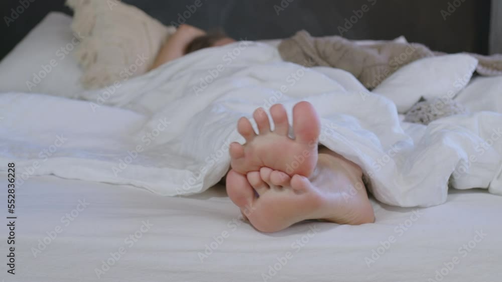 Feet peek out from under blanket of faceless girl lying in bed. Young woman moving her toes, heels, stretching after waking up in bedroom. Lady doing exercises with legs after awakening in morning Feet peek out from under blanket of faceless girl lying in bed. Young woman moving her toes, heels, stretching after waking up in bedroom. Lady doing exercises with legs after awakening in morning