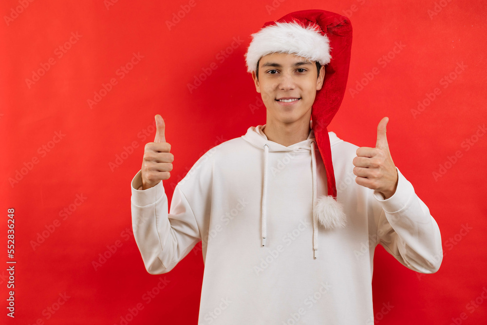 A young man, a teenager in a santa hat on a red background, points fingers up, Christmas, New Year. A young man, a teenager in a santa hat on a red background, points fingers up, Christmas, New Year.