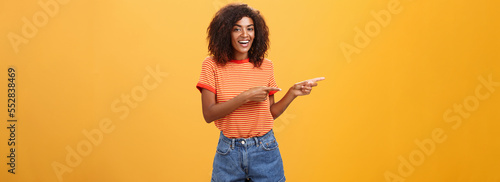 Ask him. Portrait of friendly and joyful good-looking stylish female shop assistant with curly hair and dark skin pointing left with both hands, smiling assured and entertained over orange background