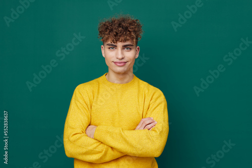 a handsome man stands on a green background in a yellow sweater and smiling pleasantly poses with his arms folded on his chest . Horizontal photo with empty space