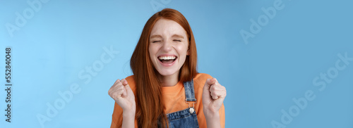 Sincere happy rejocing ginger girl close eyes smiling broadly say yes waving clenched fists joyfully celebrate enterting university dream come true winning prize triumphing cheerfully blue background photo