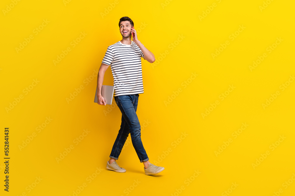 Full length photo of good mood dreamy man wear white t-shirt talking modern gadget walking empty space isolated yellow color background Full length photo of good mood dreamy man wear white t-shirt talking modern gadget walking empty space isolated yellow color background