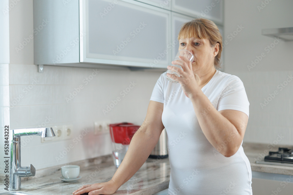 Mature woman in white t-shirt and gray leggings drinks clean water while standing in the kitchen. Mature woman in white t-shirt and gray leggings drinks clean water while standing in the kitchen.