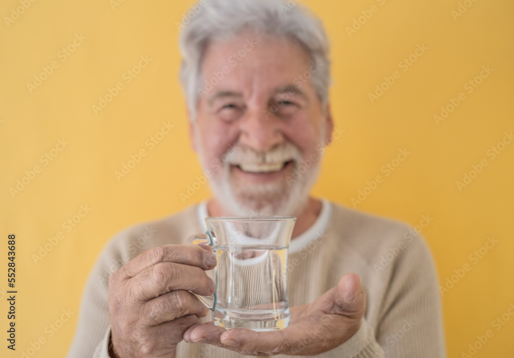 Defocused attractive bearded senior man holding a glass of fresh water looking at camera standing on yellow background Defocused attractive bearded senior man holding a glass of fresh water looking at camera standing on yellow background