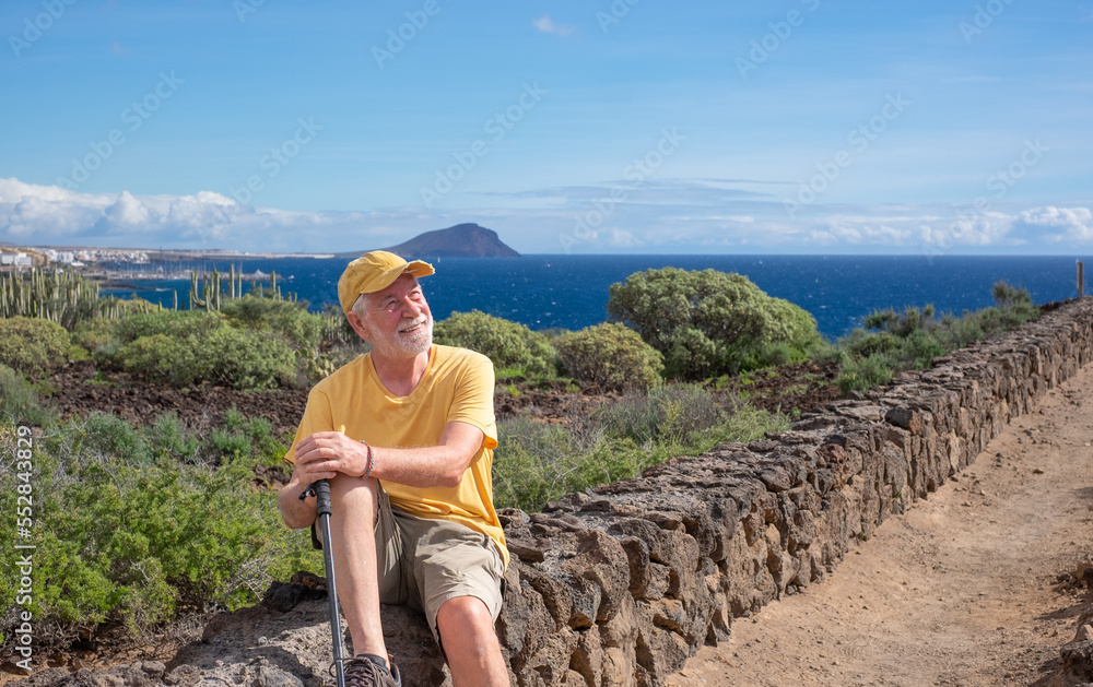 Handsome smiling senior man in hat and yellow t-shirt sitting outdoors on seaside excursion. Relaxed caucasian man enjoying his retirement by leading a healthy lifestyle