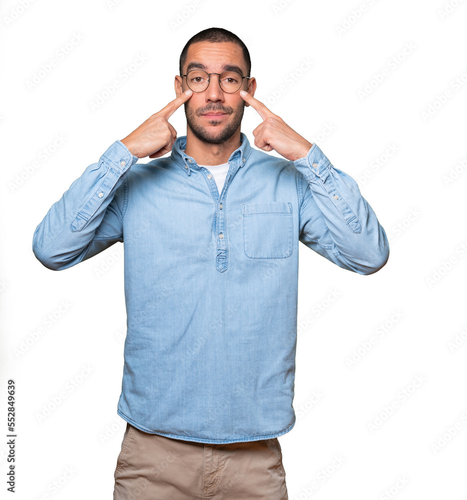 Hesitant young man making a gesture of being careful with his hand pointing at his eye Hesitant young man making a gesture of being careful with his hand pointing at his eye