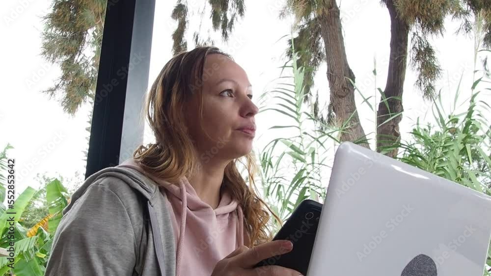 Smart modern woman sitting in a outdoor coffee shop drinking coffee and working on a laptop remotely. Distance learning online education. Happy girl makes office work remotely from cafe with computer Smart modern woman sitting in a outdoor coffee shop drinking coffee and working on a laptop remotely. Distance learning online education. Happy girl makes office work remotely from cafe with computer