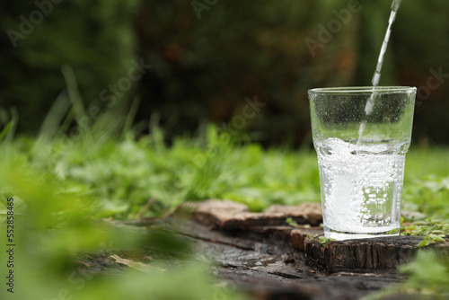 Pouring fresh water into glass on wooden stump in green grass outdoors. Space for text