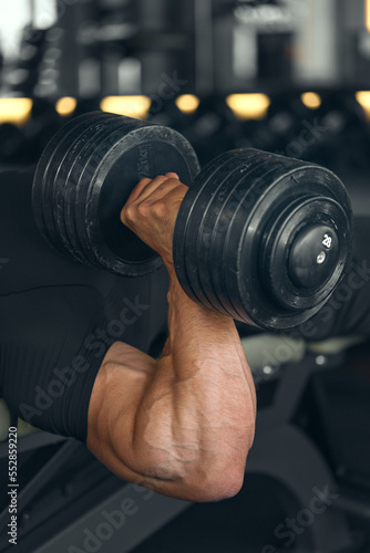 Closeup of bodybuilder hand dumbbell workout. A bodybuilder exercises with dumbbells in the gym. Young athletic and strong man training his chest muscles. Camera focus on arm