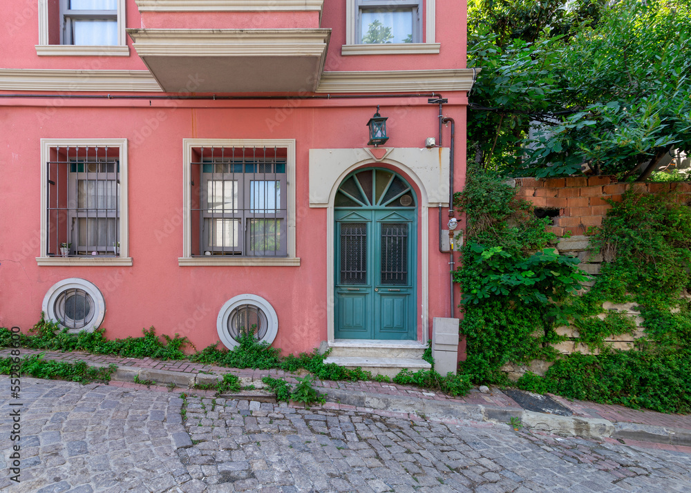Naklejka premium Exterior facade of old residential building with walls painted in pink, wrought iron windows, and green decorated door, at a narrow cobblestone street, with trees, Balat district, Istanbul, Turkey
