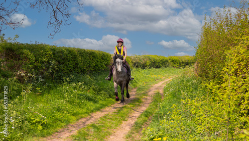 Young woman and her grey horse canter along a bridlepath on a sunny day , enjoying the freedom to move at speed and enjoy nature.