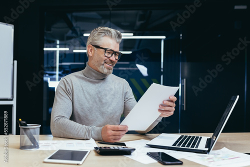 Senior successful businessman and investor working on paper work in the office, mature gray haired male boss working with contracts and accounts financier using laptop at work.