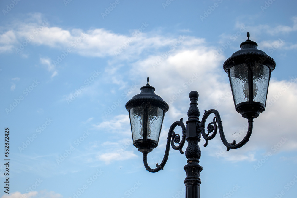 Openwork street lamps against the blue sky during the day. The outlines of ornate lighting lanterns, side view. Figured lanterns of street lighting with two plafonds.