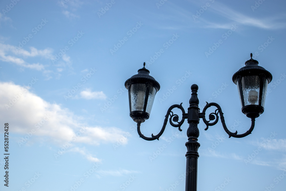 Openwork street lamps against the blue sky during the day. The outlines of ornate lighting lanterns, side view. Figured lanterns of street lighting with two plafonds.