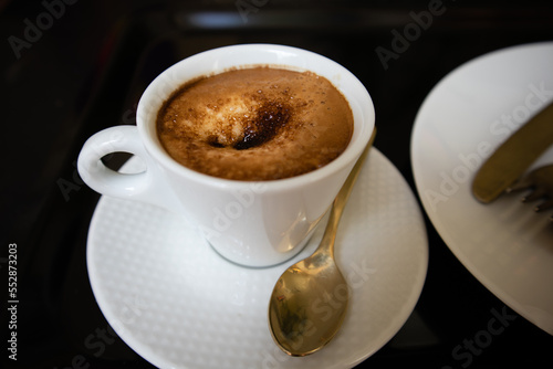 Porcelain white coffee cup with cappuccino  mochaccino on a saucer with a teaspoon on a black marble table to view. Hot tasty drink for a breakfast  branch  lunch. Aromatic beverage on a kitchen.