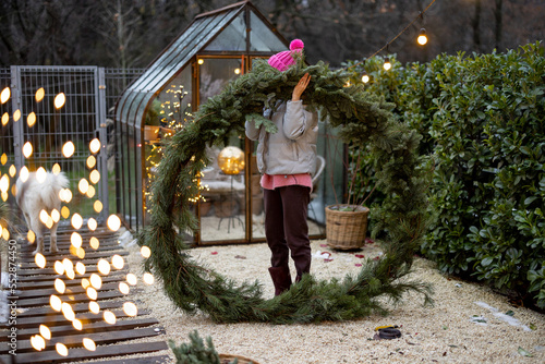 Young woman holds a big Christmas wreath made of pine twigs, preparing for a winter holidays at beautifully decorated and cozy backyard