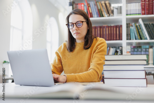 Fotografie thoughtful woman works in library or coworking at laptop and takes notes in notebook