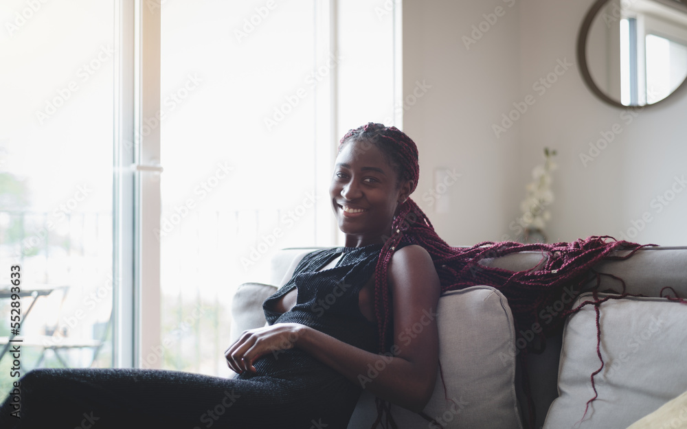 A beautiful African girl with long rose braids relaxes on the flannel fabric couch wearing a long black dress with the sunlight penetrating the windows and she is smiling for the camera A beautiful African girl with long rose braids relaxes on the flannel fabric couch wearing a long black dress with the sunlight penetrating the windows and she is smiling for the camera