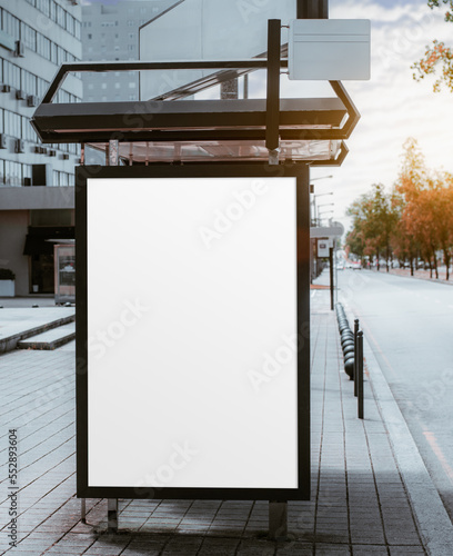 Blank vertical ad mockup in a bus terminal for commercial use, next to a residential building and with the afternoon sun flare hidden by the clouds; empty advert placeholder template on the bus stop