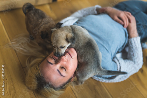 Young adult caucasian woman lying down on wooden floor with closed eyes with a brown mixed-breed puppy dog on her neck and other puppy dog standing on her blond hair. High quality photo