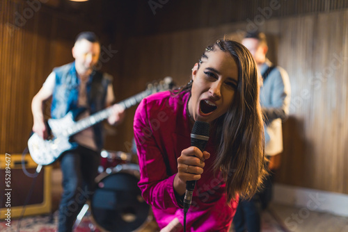 dark-haired enthusiastic woman singing into a microphone and posing for a camera her band playing in the background. High quality photo © PoppyPix