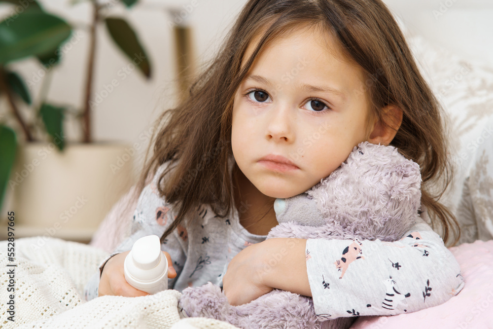 Sad depressed worried, sunless little brunette girl embracing bear toy, healing with nose spray, sit on bed and look at camera. Kid sickness, virus and covid 19. Lockdown isolation, loneliness at home Sad depressed worried, sunless little brunette girl embracing bear toy, healing with nose spray, sit on bed and look at camera. Kid sickness, virus and covid 19. Lockdown isolation, loneliness at home