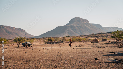 Blick auf ein Himba-Dorf in der kargen Landschaft des Kaokoveld - Im Mittelpunkt des Bildes sieht man eine Frau mit einem Wasserkanister auf dem Kopf (Kunene, Namibia) photo