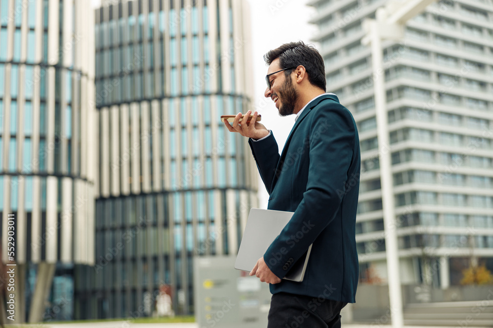 Indian Businessman with laptop recording audio message to client standing near office during break Indian Businessman with laptop recording audio message to client standing near office during break