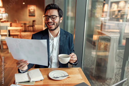 Smiling indian businessman drinking coffee and working with documents in cafe