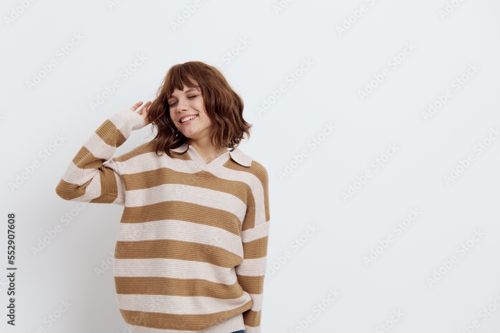 a beautiful, pleasant woman stands in a stylish striped sweater with her hands raised behind her head on a light background, closing her eyes and smiling broadly a beautiful, pleasant woman stands in a stylish striped sweater with her hands raised behind her head on a light background, closing her eyes and smiling broadly