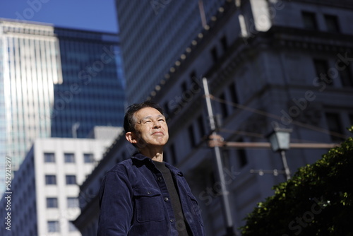 Middle-aged Asian man looking up at blue morning sky with high-rise business building in background.