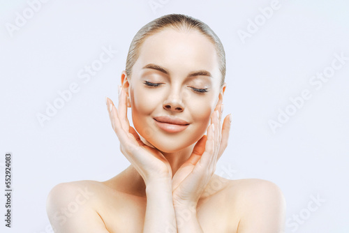 Close up portrait of young beautiful girl closes eyes  touches face gently  has well cared complexion  healthy skin  enjoys facial treatment  stands shirtless against white wall. Freshness  body care