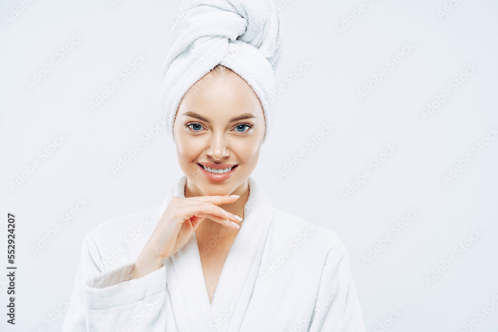 Women, beauty, hygiene, wellness concept. Fresh young European woman wears bath towel and robe, touches chin gently, spends free time in spa, undergoes beauty treatments after shower white background. Women, beauty, hygiene, wellness concept. Fresh young European woman wears bath towel and robe, touches chin gently, spends free time in spa, undergoes beauty treatments after shower white background.
