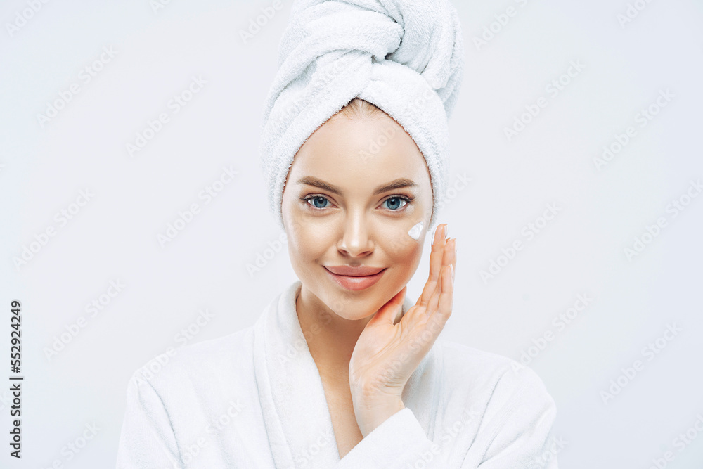 Young confident Caucasian woman applies face cream, enjoys new anti wrinkle cosmetic product, prevents sign of skin aging, wears minimal makeup, dressed in bath robe, isolated on white wall. Young confident Caucasian woman applies face cream, enjoys new anti wrinkle cosmetic product, prevents sign of skin aging, wears minimal makeup, dressed in bath robe, isolated on white wall.