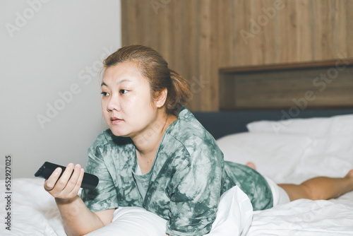 Obraz na plátně Asian housewife lying prone in bed and holding remote control watching television with relaxing action
