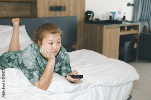 Fotografie Asian housewife lying prone in bed and holding remote control watching television with relaxing action
