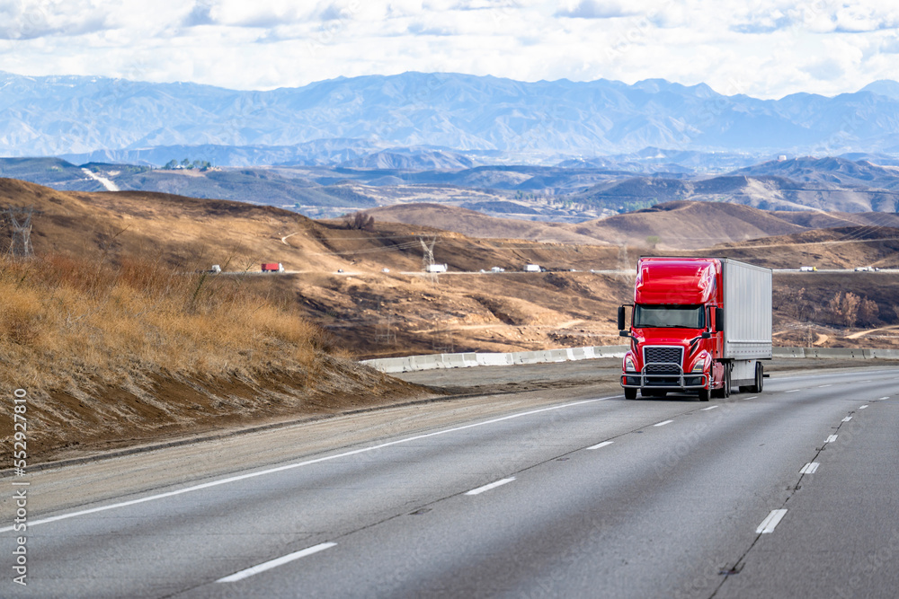 Bright red big rig bonnet semi truck transporting cargo in dry van semi trailer slowly climbing uphill on the mountain highway road Bright red big rig bonnet semi truck transporting cargo in dry van semi trailer slowly climbing uphill on the mountain highway road