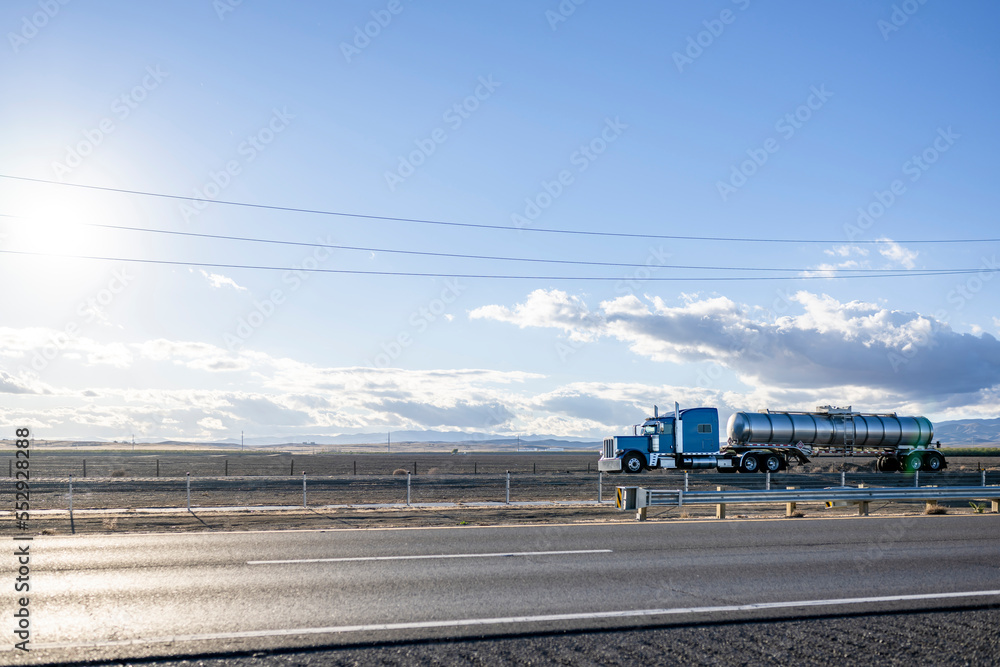 Naklejka premium Classic powerful blue big rig semi truck with chrome accessories transporting liquid cargo in stainless steel tank semi trailer driving on the straight highway road between fields