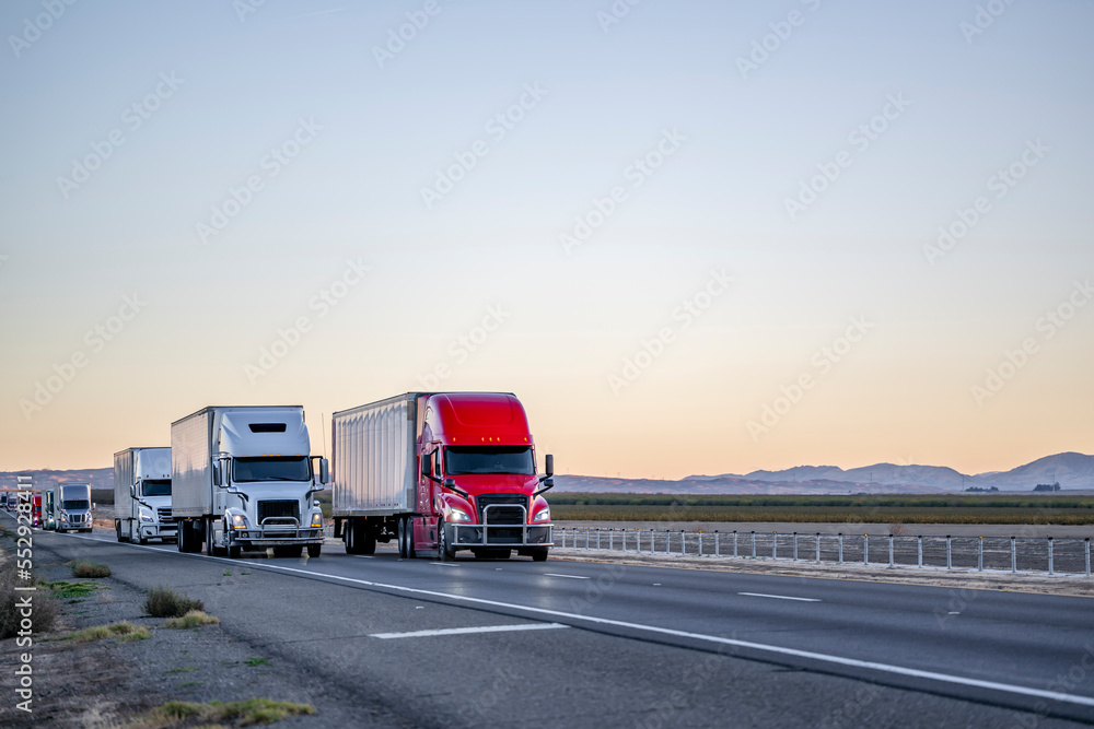 Convoy of industrial big rigs semi trucks with semi trailers driving with cargo on the straight highway road at twilight time