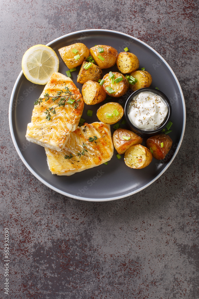 grilled cod fish with baked jacket potatoes, cream sauce and lemon close-up in a plate on the table. Vertical top view from above grilled cod fish with baked jacket potatoes, cream sauce and lemon close-up in a plate on the table. Vertical top view from above